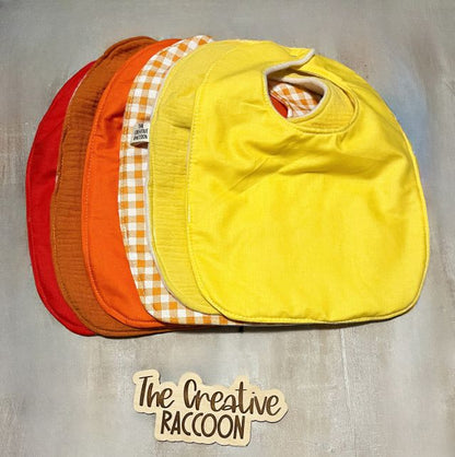 Four colorful bibs stacked on a textured surface with 'The Creative Raccoon' branding. Solid colored baby bibs - red, burnt orange, orange, yellow, and sunshine yellow. Patterned baby bibs - orange and white gingham. Other bibs are available. Handmade in Kansas, USA
Football Baby Gift Sets, Kansas Theme - The Creative Raccoon 