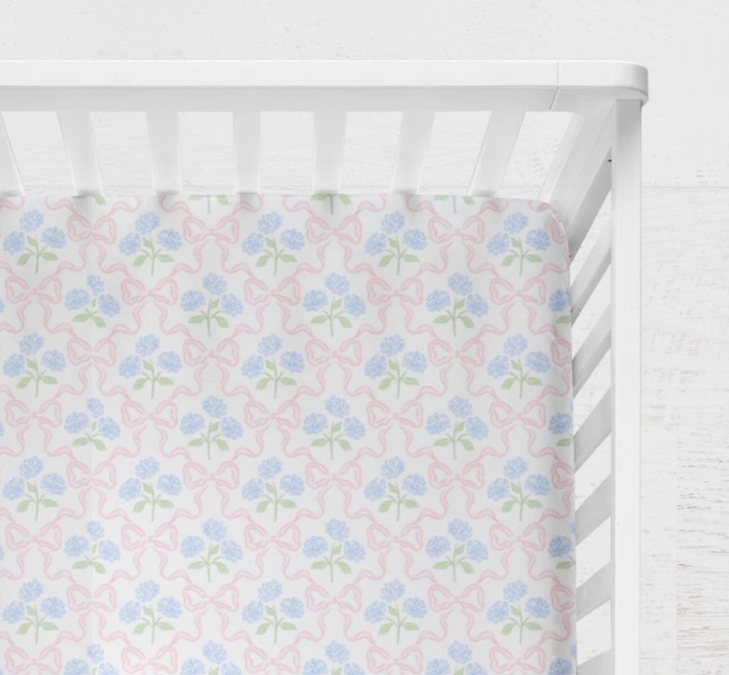 Floral-patterned crib sheet with pink and blue Hydrangeas and Bows flowers on a white crib. Hydrangeas and Bows Crib Sheet - The Creative Raccoon