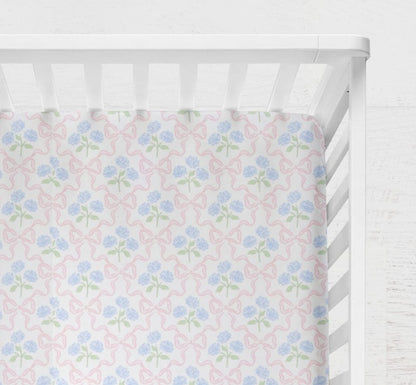 Floral-patterned crib sheet with pink and blue Hydrangeas and Bows flowers on a white crib. Hydrangeas and Bows Crib Sheet - The Creative Raccoon