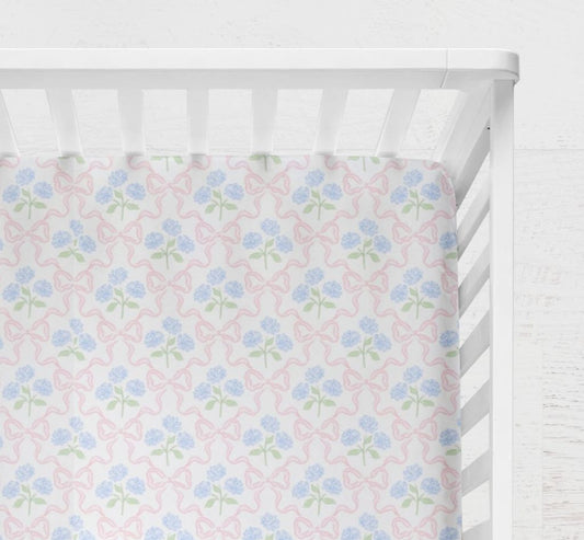 Floral-patterned crib sheet with pink and blue Hydrangeas and Bows flowers on a white crib. Hydrangeas and Bows Crib Sheet - The Creative Raccoon