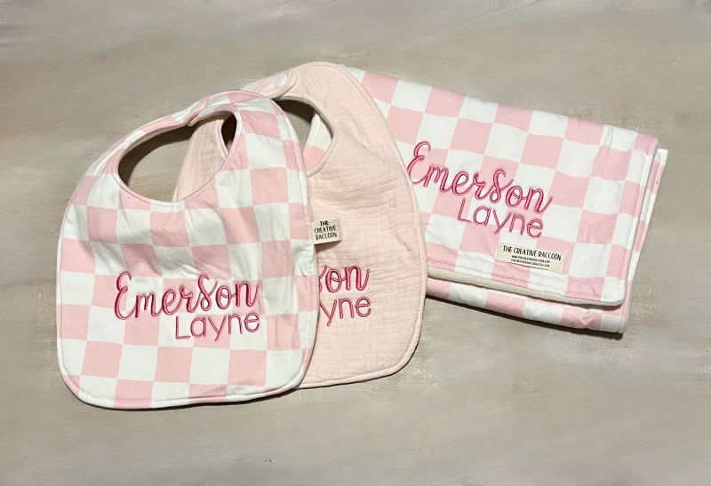 Pink checkered baby bib and burp cloth with 'Emerson Layne' embroidered on both items on a beige surface. Pink Checkered Baby Bib and Burp Cloth Personalized