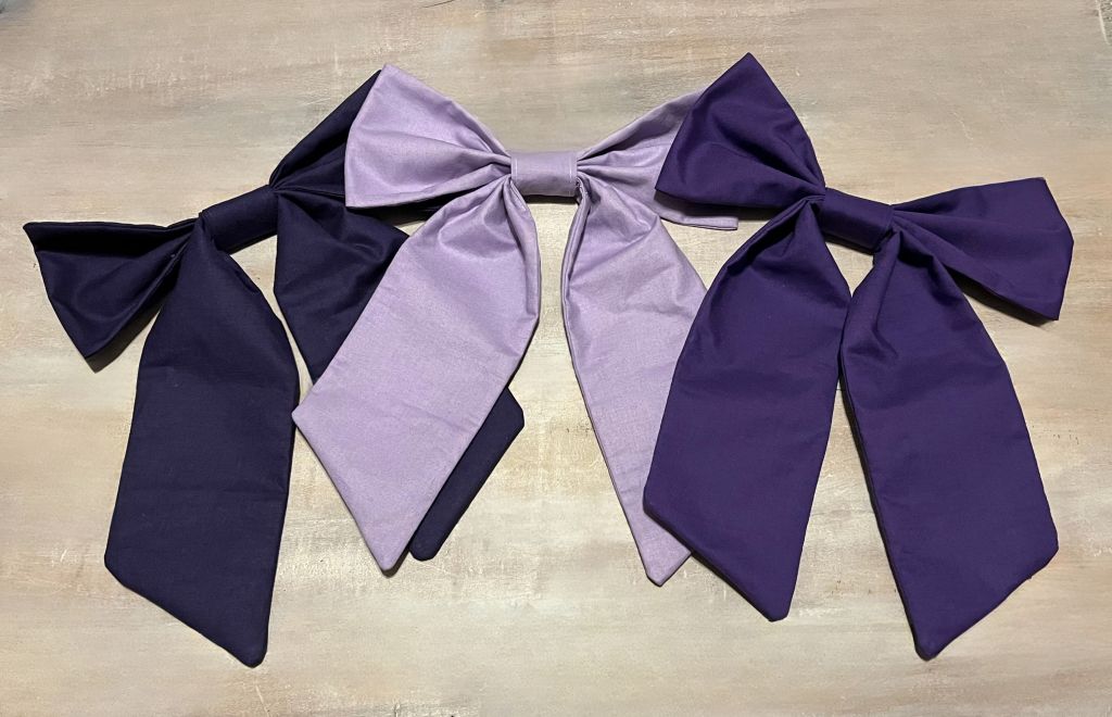 Three large fabric bows in purple and lavender on a wooden surface. Purple bow add-ons for car seat canopy or crib decor. - The Creative Raccoon
