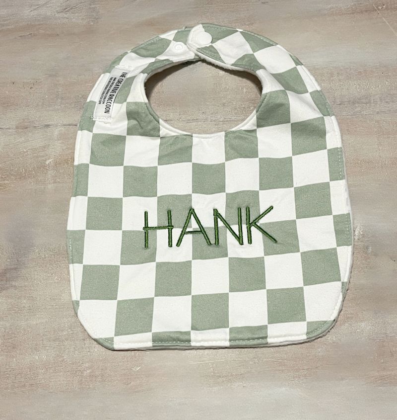 Green and white checkered bib with 'HANK' embroidery on a wooden 
surface.
Sage Green Checkered Bib and Burp Cloth Set - the Creative Raccoon