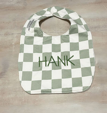 Green and white checkered bib with 'HANK' embroidery on a wooden 
surface.
Sage Green Checkered Bib and Burp Cloth Set - the Creative Raccoon