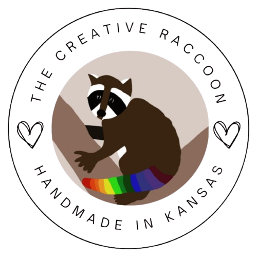 Logo of 'The Creative Raccoon' with a raccoon a rainbow towel, surrounded by hearts and text on a white background.