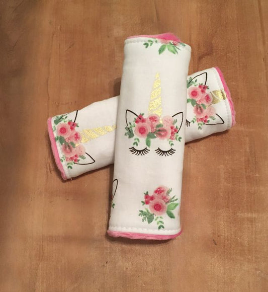 Two unicorn-themed car seat belt covers with floral designs on a wooden surface. Unicorn Themed Car Seat Strap Covers - The Creative Raccoon