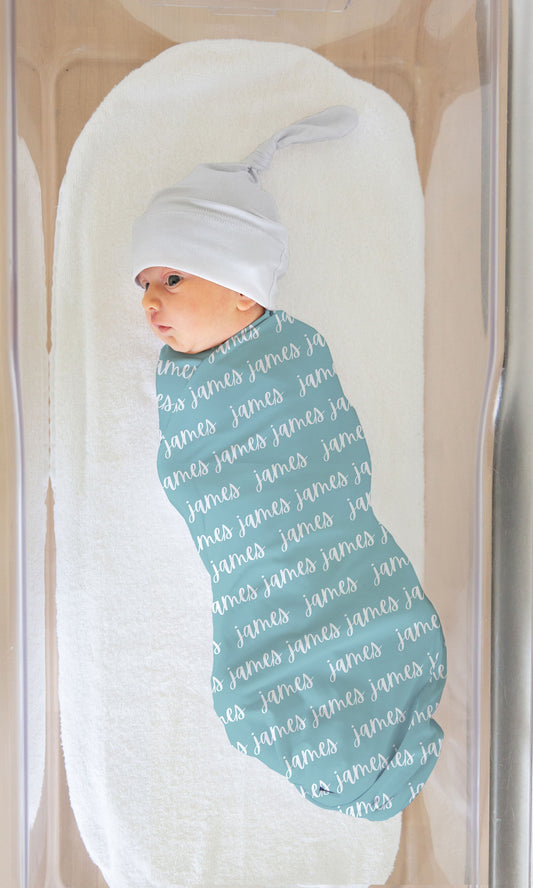 aqua blue swaddle blanket with custom name in white, made out of jersey knit. The design is printed on one side. Blanket shown on a baby in a bassinet